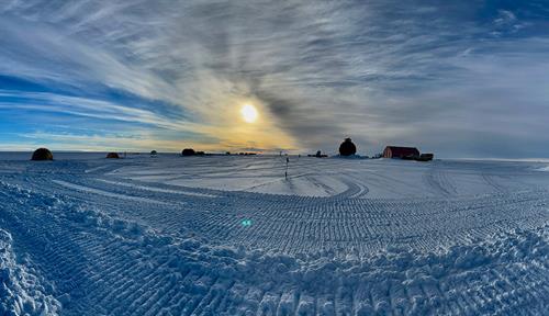Fotografía de un paisaje llano y desolado, cubierto de hielo y nieve, con el sol bajo en el cielo. En el fondo se ven algunas estructuras.