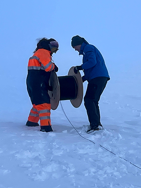 Fotografía de dos trabajadores abrigados que sostienen un gran carrete y desenrollan el cable en la nieve.