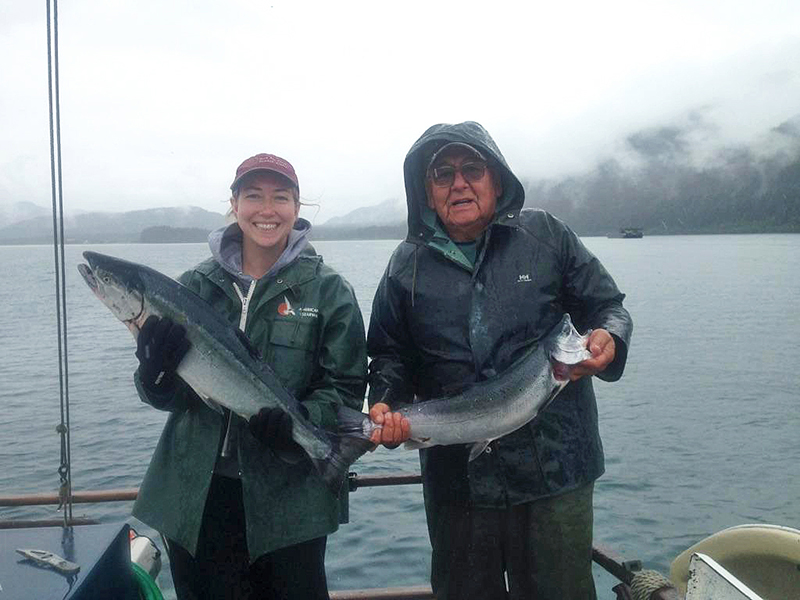 Una mujer y un hombre posan sosteniendo cada uno un salmón que han pescado. Al fondo se ve agua y una montaña cubierta de nubes.