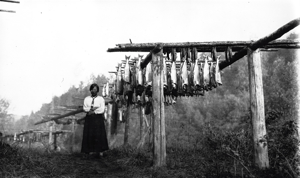 Una imagen en blanco y negro capta a una mujer junto a un secadero de salmones con docenas de peces.
