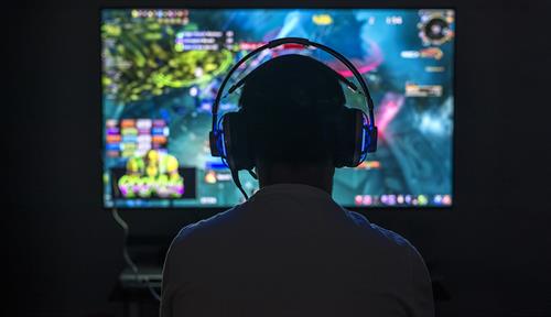 Silueta de jugador de videojuegos visto de espaldas, con auriculares. Delante del jugador hay una pantalla de computadora de colores brillantes.