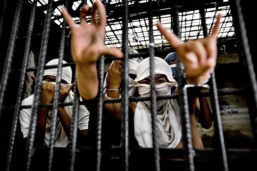 Jóvenes dentro de una celda de prisión se muestran con sus rostros tapados con tela blanca. Uno de ellos, en primer plano, realiza señales con sus manos.