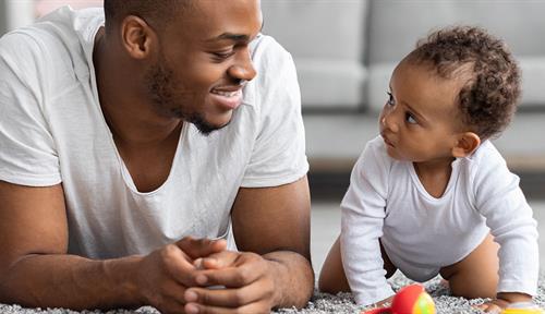 La foto muestra a un joven negro sonriendo a un bebé que gatea en un salón.