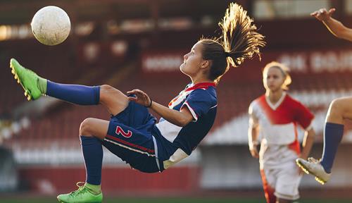 Una adolescente salta en el aire para patear una pelota de fútbol mientras otras jugadoras observan.