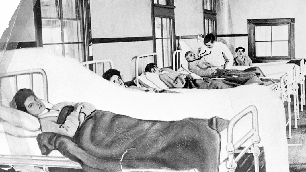 Foto de una mujer acostada en una cama de un hospital. Otras mujeres yacen en camas al lado de ella. La mujer en primer plano tiene los brazos cruzados y mira directo a la cámara.