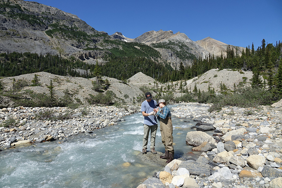 Foto de dos personas de pie sobre una gran roca en un arroyo, examinando una muestra. Los rodean las montañas rocosas.