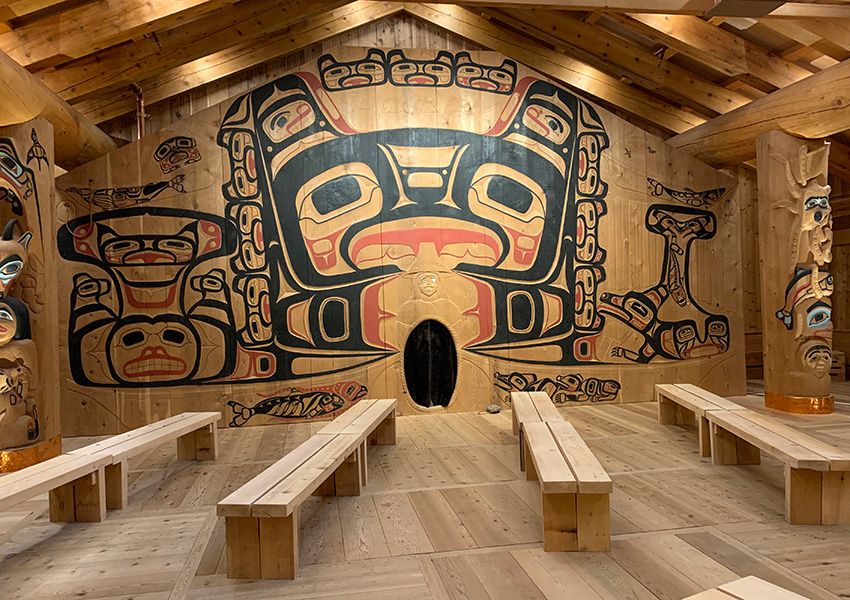 La foto muestra el interior de una cabaña de madera con tallas, murales brillantes pintados en las paredes y una serie de bancos de madera.
