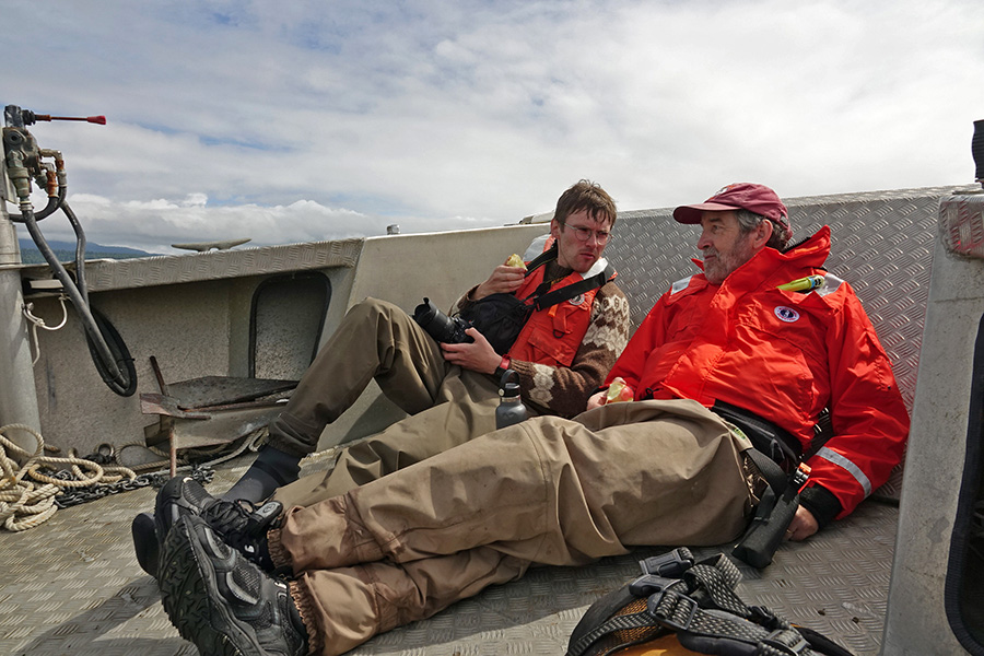 Dos hombres descansan en la cubierta de un barco comiendo manzanas y hablando. Llevan botas de montaña y ropa de abrigo. El cielo está nublado.