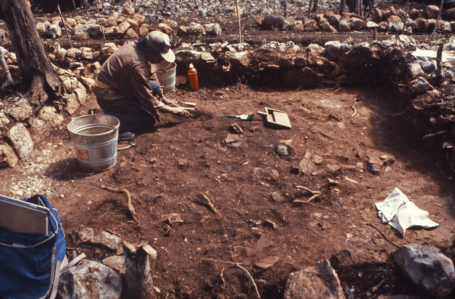 Fotografía de un hombre agachado retirando polvo con una brocha. Está rodeado por ruinas de muros de piedra. En las cercanías se observan baldes y otras herramientas para excavación arqueológica. 