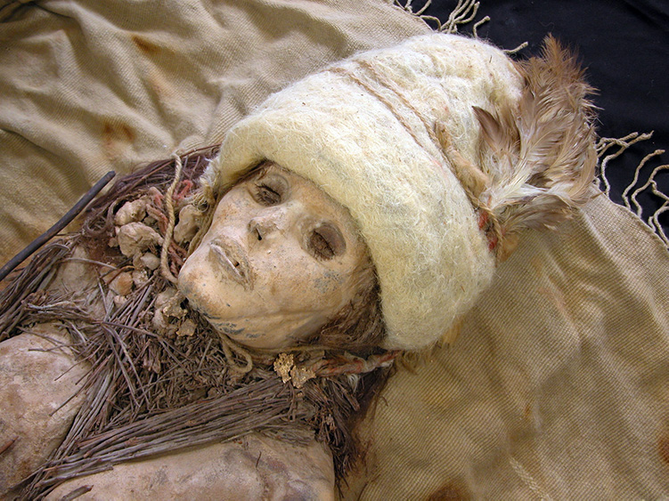 Foto de primer plano de la cabeza y los hombros de una momia muestra un esqueleto que lleva un sombrero con una pluma, una cuerda alrededor del cuello con pequeños mechones atados a ella y ramitas.