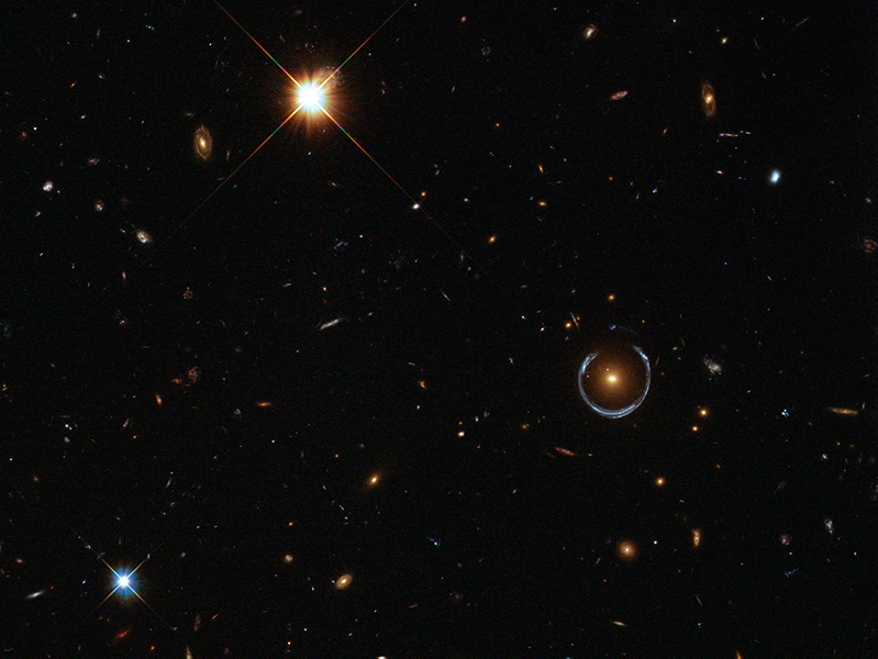 La imagen de Hubble muestra una vista profunda del espacio con un semicírculo azulado en forma de herradura alrededor de un objeto brillante que se ve entre las estrellas y las galaxias. Los científicos creen que la herradura es una galaxia con un desplazamiento al rojo de aproximadamente 2,4, lo que significa que la vemos tal y como era cuando el universo tenía apenas 3.000 millones de años.