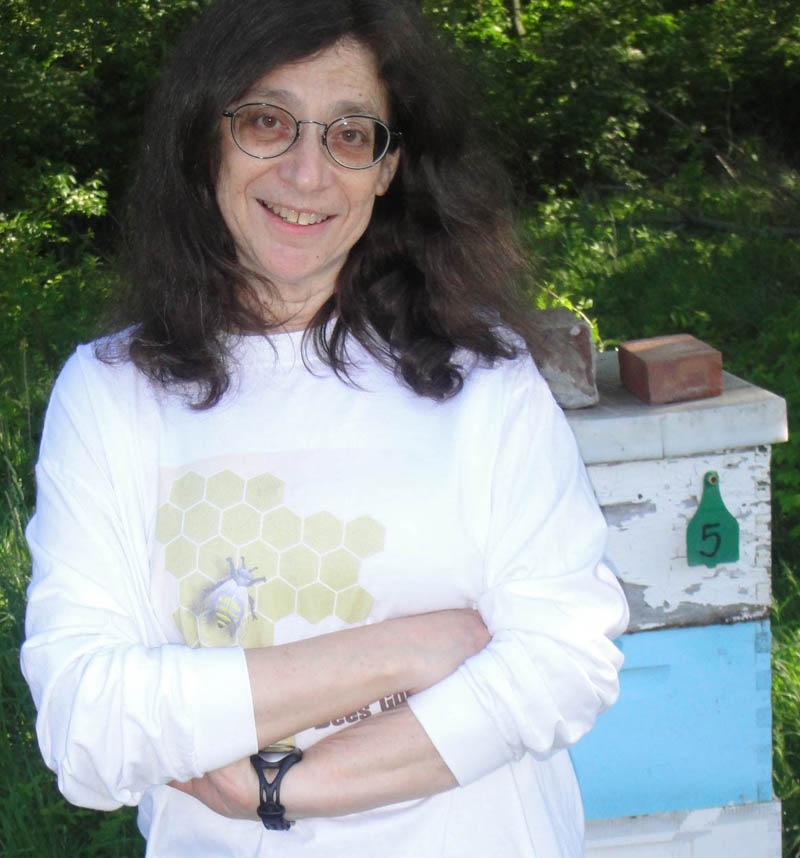 Fotografía de May Berenbaum vistiendo una camiseta con un panal, frente a un colmenar. 