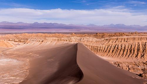 Una enorme duna de arena pardusca, rocas escarpadas de color marrón anaranjado detrás y montañas violáceas a lo lejos.