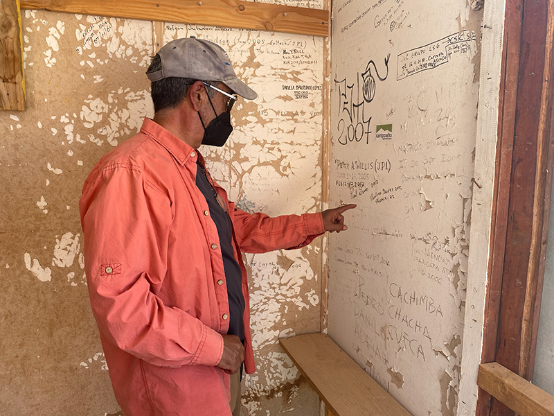 Un hombre señala una pared con pintura desconchada; los nombres de las personas están garabateados por toda la pared