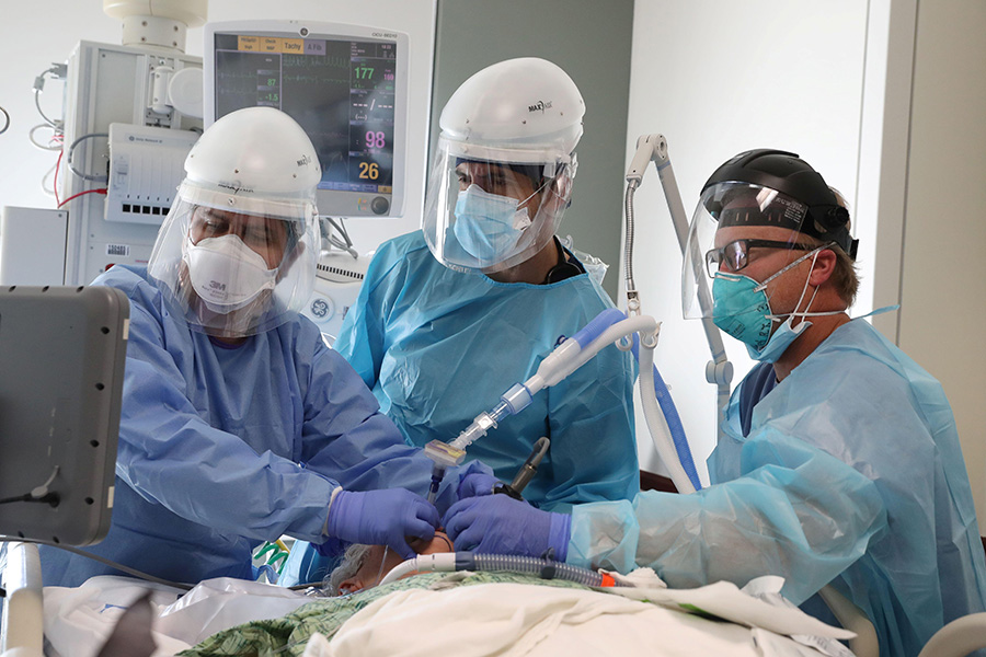  Texto alternativo de la foto: Una fotografía muestra a tres trabajadores sanitarios vestidos con mascarillas, protectores faciales, batas y guantes alrededor de la cama de un paciente en la unidad de cuidados intensivos. Están intubando al paciente y observando el progreso de la intubación en una pantalla.