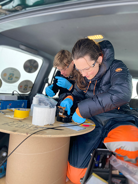 Photograph of a woman and a man, both wearing blue gloves and padded clothing, sitting in a vehicle splicing cables. There is a large cable spool in the foreground that they’re using as a table for their work.
