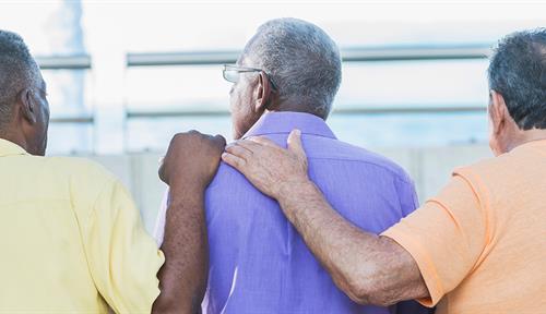 Photos shows three older men sitting on a park bench, with two of them comforting the man in the middle.