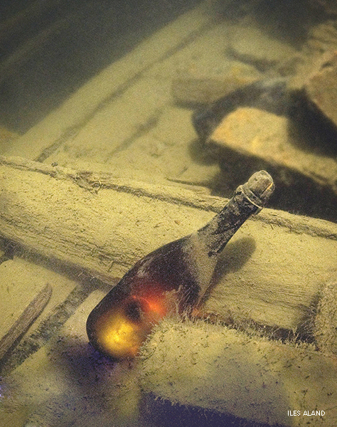 Fotografía de una botella de champán cubierta de sedimentos, bajo el agua. Se encuentra entre los restos de un barco.