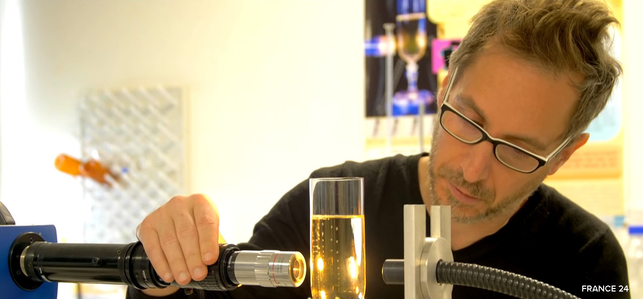 Fotografía de un hombre con gafas y barba y bigote claros que lleva un suéter negro; está inclinado sobre una cámara de alta velocidad que está dirigida a una copa de champán para poder estudiar las burbujas que se están formando.