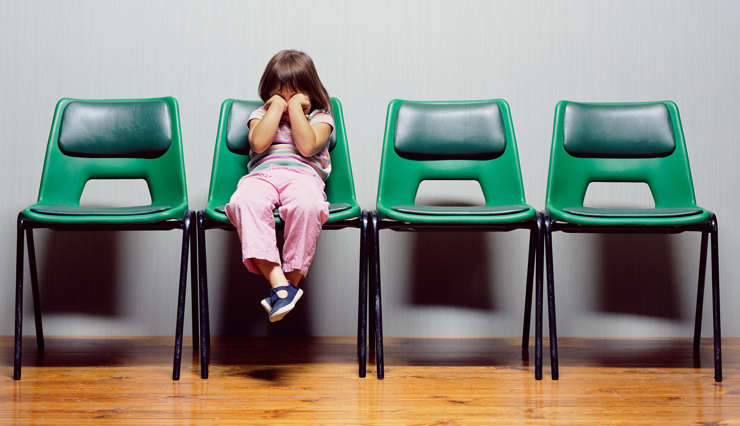 Fotografía de una niña pequeña sentada en una silla de la sala de espera con los puños tapándole la cara.