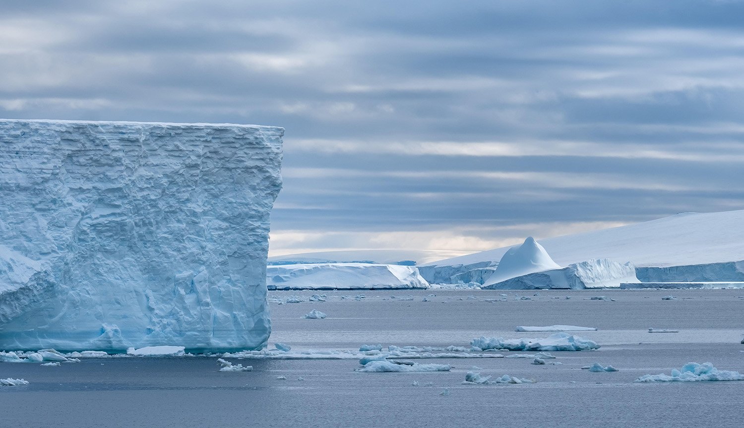 Un vasto acantilado de hielo se eleva sobre aguas abiertas, con icebergs y glaciares al fondo, cerca de la plataforma de hielo de Ross, en la Antártida.