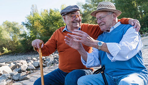 Photograph of two elderly men sitting outdoors. They are enjoying a good joke or tale. The man on the left has his arms around the shoulders of his friend. He ls laughing. The man on the right has his arms out in front, slightly raised, as if he is relating a story. He is grinning.
