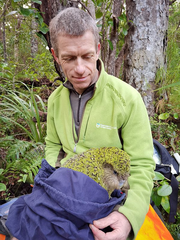 El biólogo conservacionista Andrew Digby sentado en un bosque sosteniendo un kākāpō envuelto en tela azul.