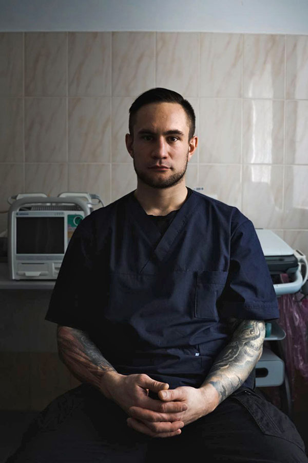 Cirujano ucraniano Un joven serio con bata azul oscuro, con los brazos tatuados cruzados delante de él, en una habitación de hospital.