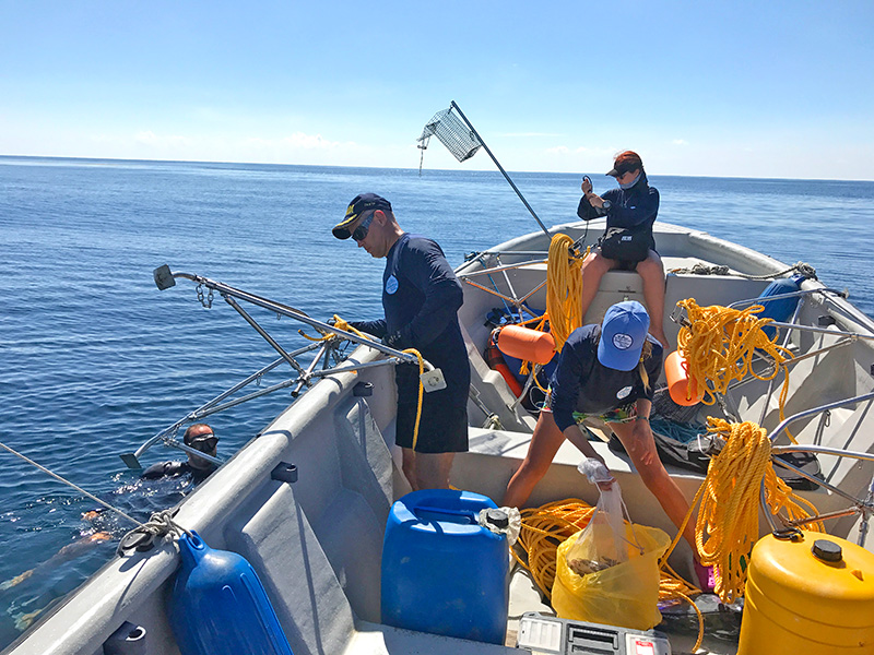 En medio del océano, se observan tres personas a bordo de una lancha y otra persona en el agua. En el bote, hay recipientes azules y amarillos, así como cables y otros materiales.