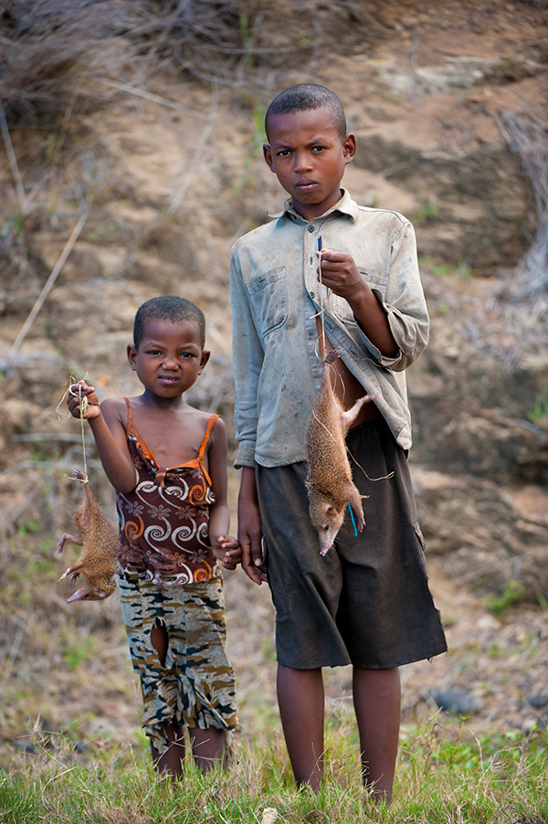 Fotografía de dos niños de pie en la hierba, con rocas y tierra detrás de ellos. Cada uno de ellos sostiene un mamífero muerto de color marrón y hocico largo atado a un trozo de cordel.