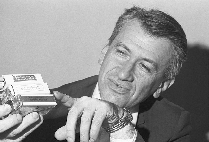 Fotografía en blanco y negro de un hombre señalando una etiqueta de advertencia en un paquete de cigarrillos.