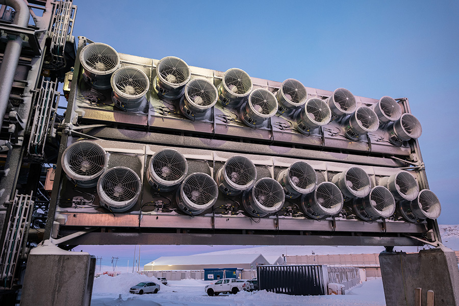 La foto muestra una fila de una docena de ventiladores circulares en cada uno de los dos contenedores metálicos colectores de dióxido de carbono, con el cielo azul y un aparcamiento nevado y edificios al fondo.