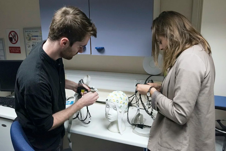 Gorro EEG Fotografía de dos personas trabajando con equipos electrónicos en una mesa de laboratorio. Están inclinados sobre un modelo de cabeza humana sobre la que se ha colocado un gorro con electrodos.