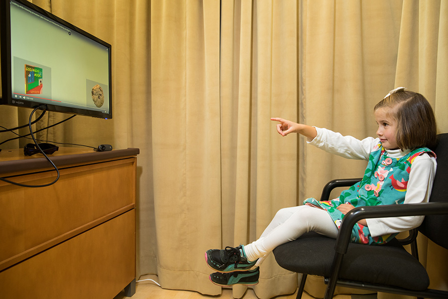 Una niña está sentada en una silla de oficina y señala las imágenes que está viendo en un monitor frente a ella.