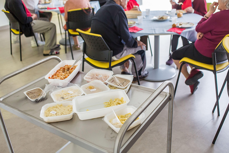 Foto de alimentos en cajas de cartón sobre una bandeja; cerca hay mesas con residentes de hogares de ancianos sentados a su alrededor. Los alimentos son insípidos en tonos marrones y beige.