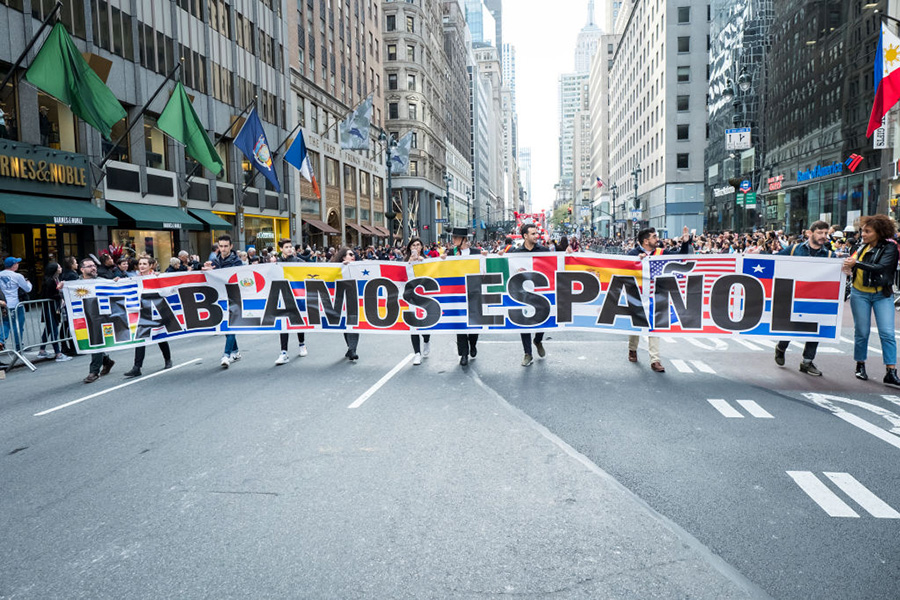 Personas marchan en medio de una avenida portando un cartel con banderas de países hispanos y escrita sobre ellas la frase 