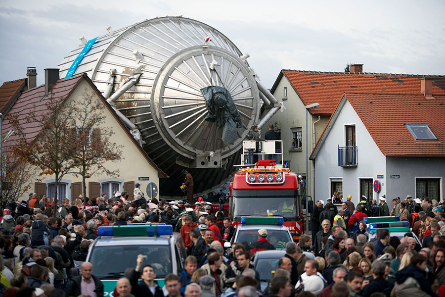 La foto muestra el transporte del enorme espectrómetro metálico KATRIN por una calle estrecha, sobresaliendo por encima de las casas a ambos lados, en el centro de Leopoldshafen, Alemania, con gente reunida para presenciar el evento.