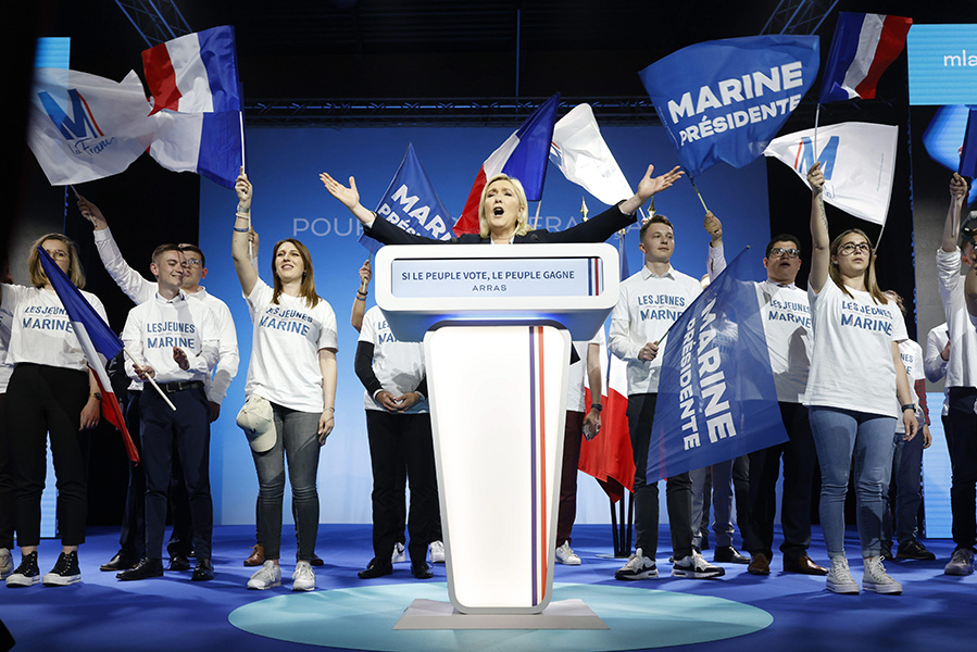 Una mujer rubia frente a un atril con los brazos en alto mientras gente con camisetas de “Marine” ondea banderas francesas y pide que sea presidenta. En el atril está escrito “si le peuple vote, le peuple gagne” (si el pueblo vota, el pueblo gana).