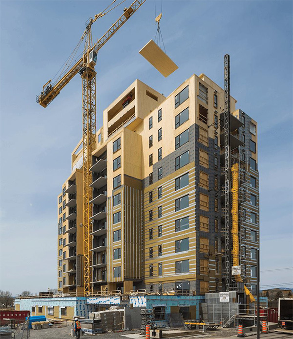 Una foto muestra un edificio alto de madera en masa en construcción, con una grúa bajando un gran panel de madera en su lugar encima de la estructura.