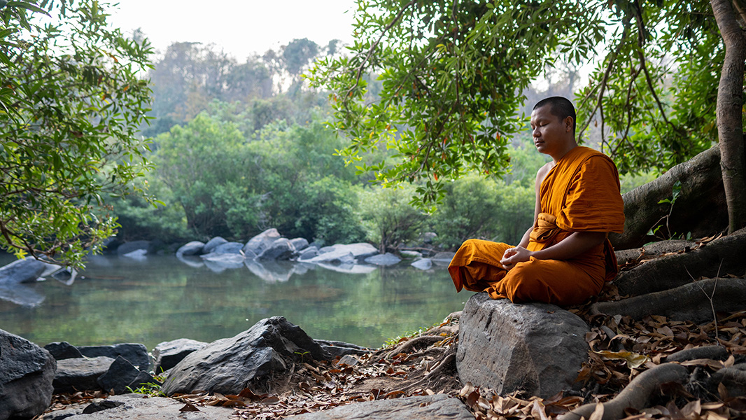 Un hombre vestido de naranja está sentado en una roca con los ojos cerrados, meditando. Al fondo, un río, rocas y árboles.