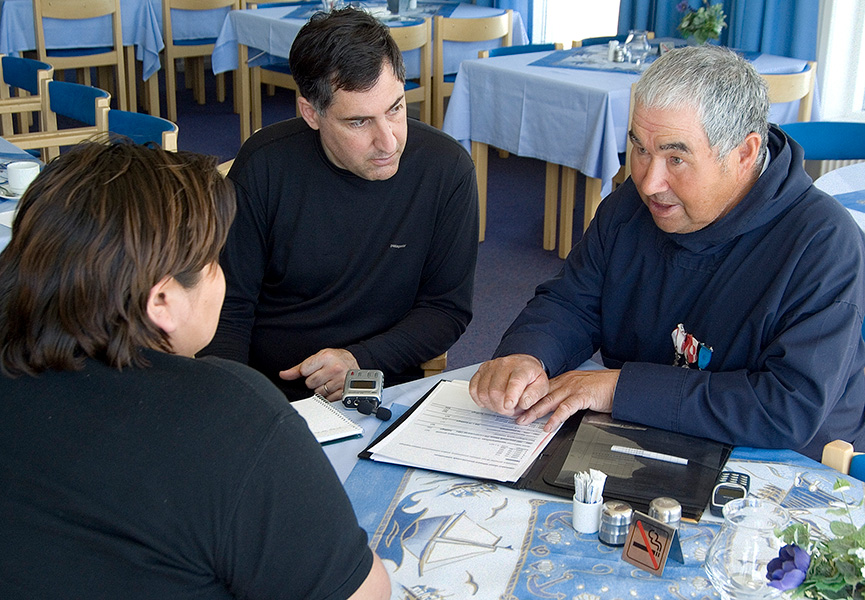 Tres personas están sentadas en una mesa. Una de ellas, un hombre mayor, está hablando y consultando un cuaderno, mientras las otras dos escuchan atentamente.