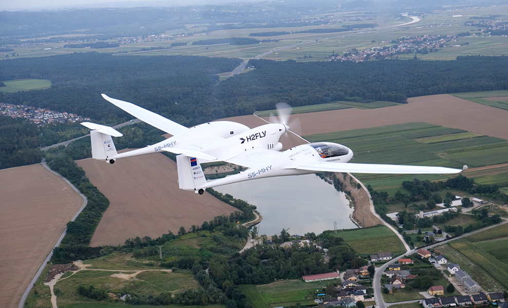 Un pequeño avión blanco con la inscripción “H2FLY” en el costado vuela sobre campos y casas.