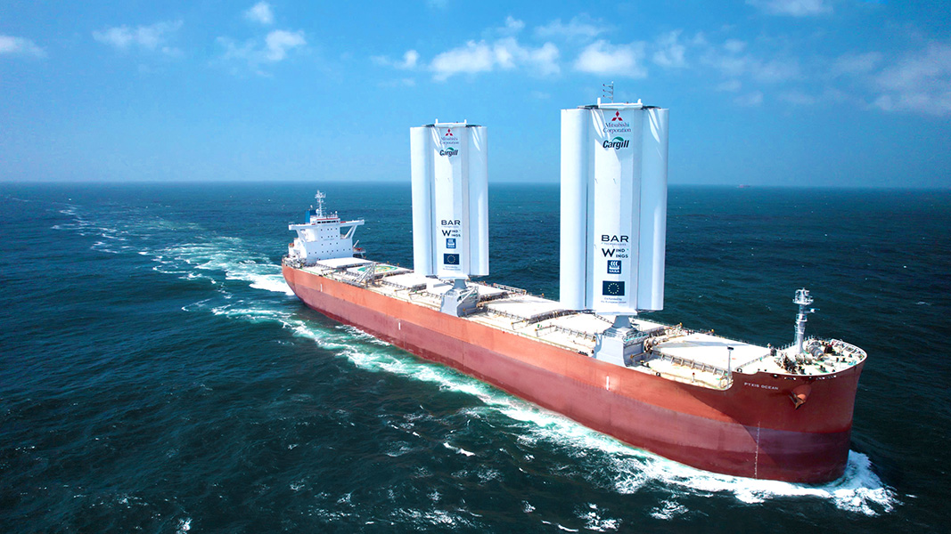 Foto de un enorme barco en el agua, con el casco rojo óxido y dos grandes estructuras —las velas— que parecen varios cilindros verticales unidos entre sí. 