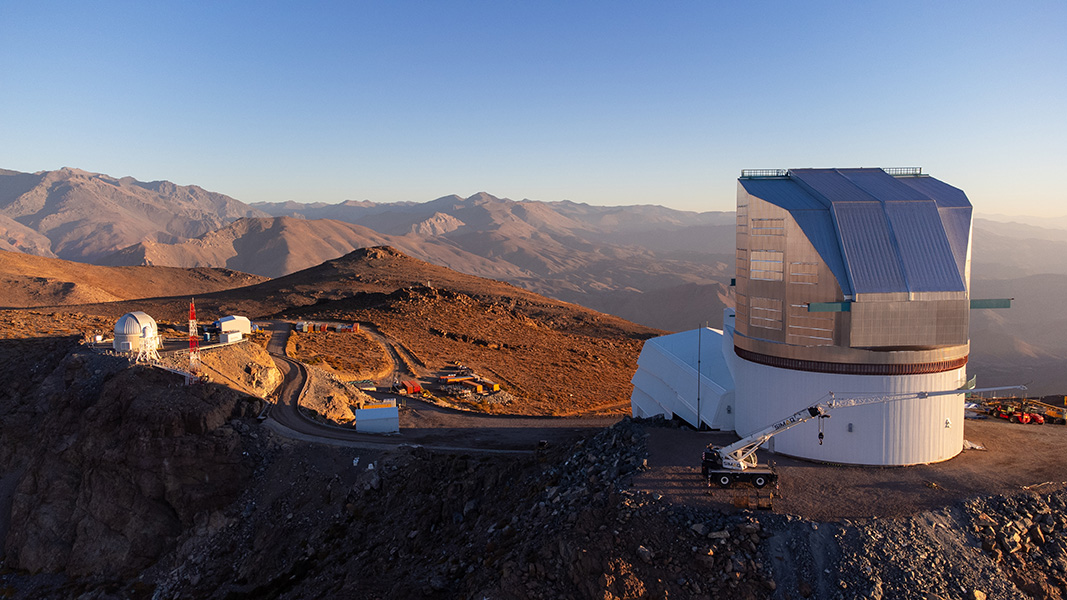 Vista al atardecer del Observatorio Rubin en lo alto de su emplazamiento en la montaña desértica chilena; las crestas montañosas se alejan en la distancia bajo un cielo azul.