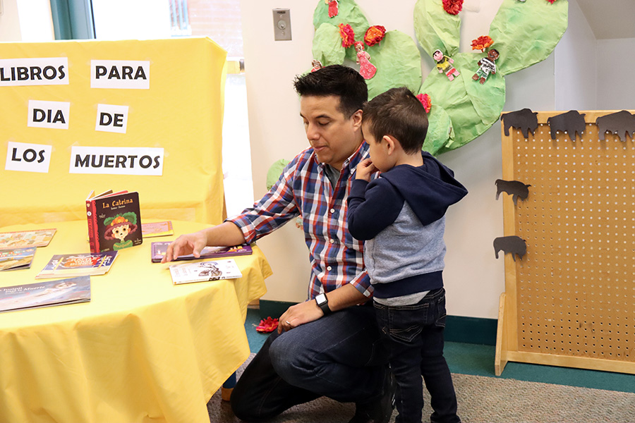 Un niño junto a un hombre arrodillado ante una mesa con libros infantiles. Al fondo hay un cartel que dice “Libros para día de los muertos” 