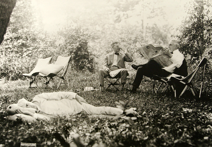 Una fotografía histórica muestra a un hombre con traje blanco durmiendo en el césped mientras otros dos hombres están sentados en tumbonas detrás de él leyendo periódicos.