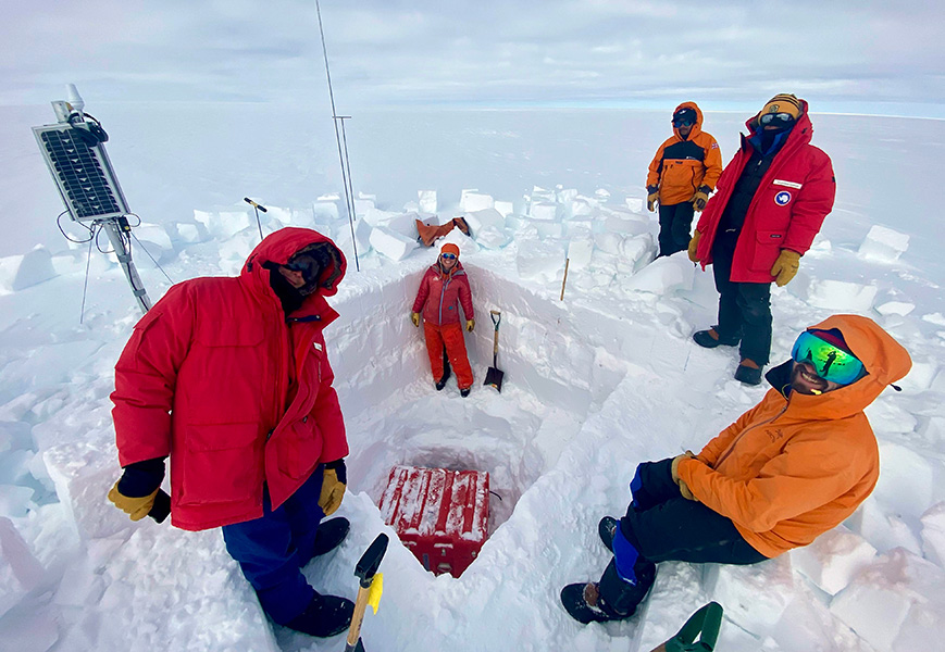Varias personas con ropa para el frío posan cerca de un agujero que han cavado en un glaciar, con varias piezas de equipo cerca.