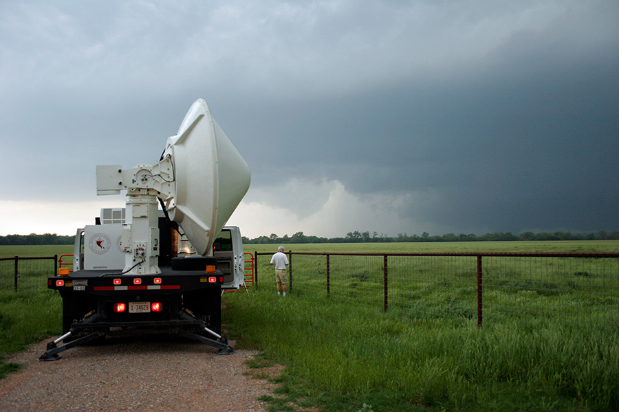 Un camión visto desde la parte trasera con un artilugio de radar gigante en su cama, una persona se encuentra cerca mirando a las nubes altas engendrando un tornado en la distancia.