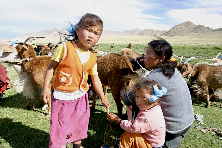Foto de una mujer en cuclillas en el suelo, con dos niños delante y varias cabras en el fondo.
